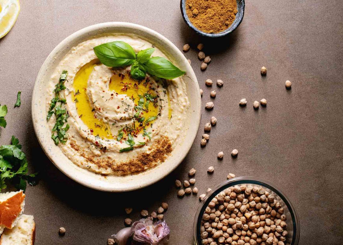 Hummus with olive oil and ground cumin in ceramic bowl served with lemons, basil and chickpeas over brown texture background. Top view, flat lay